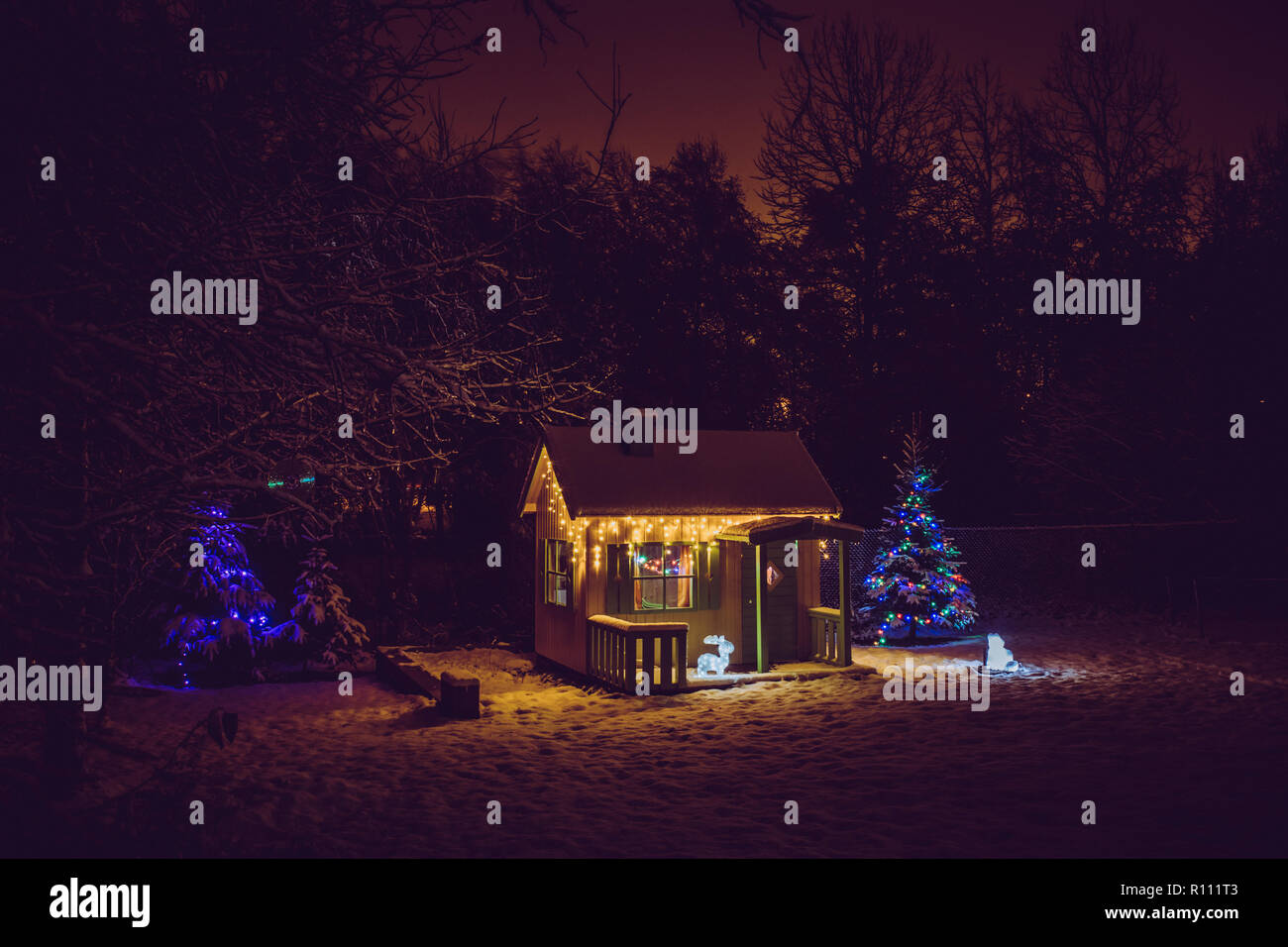 Wooden painted yellow private children`s play house in home garden, decorated with Christmas LED string lights outdoors in cold winder night. Decorate Stock Photo